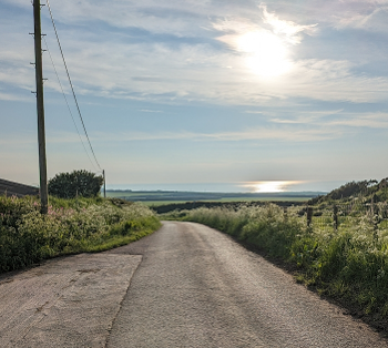 A road turns down a hill in countryside, with shining sea at horizon and sparse clouds.