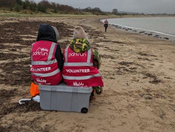 Two parkrun volunteers sit on a crate on a beach, watching the runners approach.