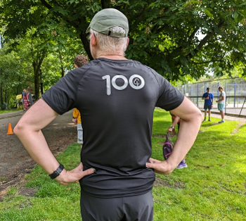 A photo of the back of a 100 tee shirt, with a guy in black shorts wearing it.