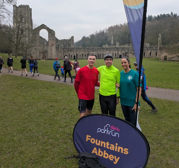 A group of three in front of the parkrun sign at Fountains Abbey, with the abbey behind.