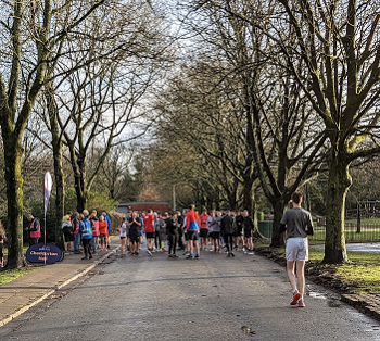 Parkrunners assemble on wide tarmac path in a tree-lined park.