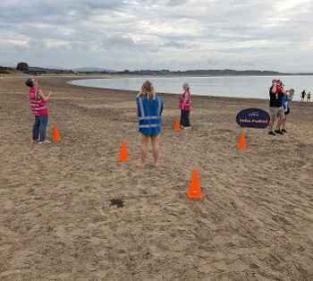 The finish funnel at a parkrun. Orange cones on sand. Marshals in fluorescent jackets.