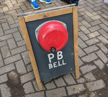 A red crank bell, mounted on a sandwich board, with the caption PB Bell.