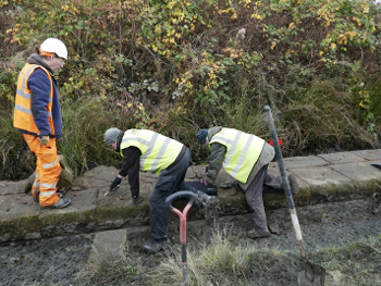 2 people half kneeling excavating, being watched by someone in orange