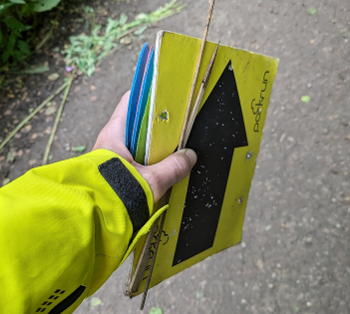 A yellow-sleeved hand holding a parkrun arrow board, stakes and stacked plastic cones.