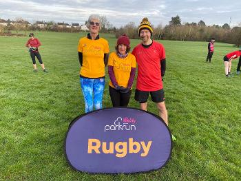 3 runners behind the Rugby parkrun sign